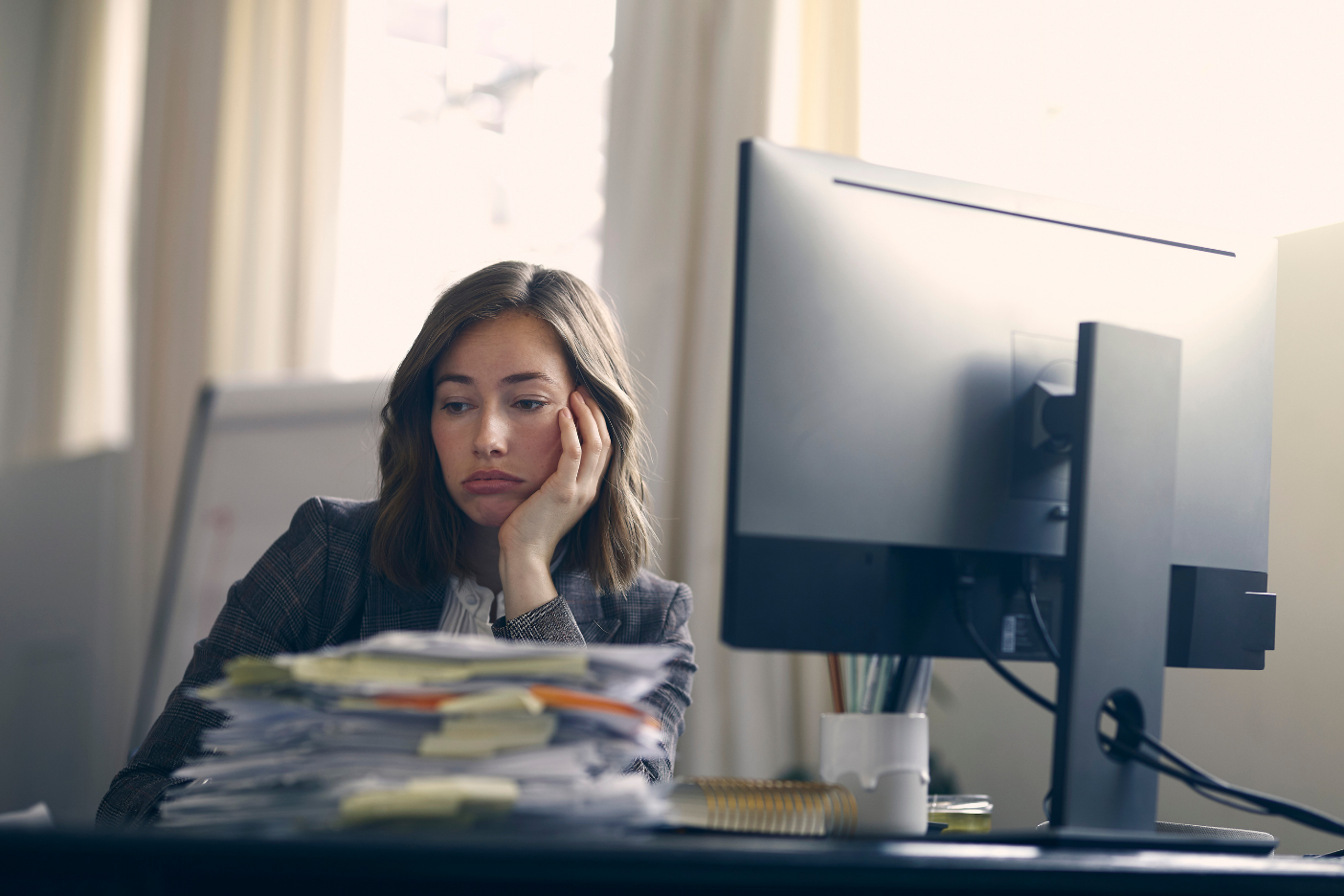 Unhappy business woman is tired of her job. Sitting infront of her computer with a large pile of paperwork on her desk.